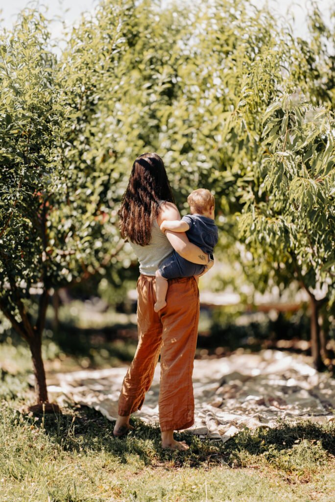 Photograph of the blog author, Hannah Yinhar, a therapist and mother, holding her son in an orchard with their backs to the camera during postpartum. A photo of what motherhood is like.